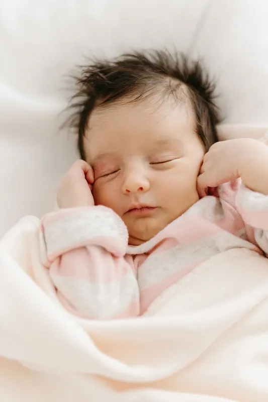 Newborn baby with dark hair sleeping peacefully, tucked under a soft pink blanket