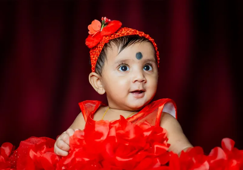 Wide-eyed baby girl in a red frilly dress and flower headband against dark curtains