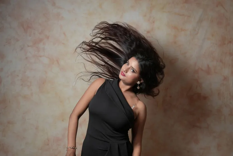 Model in a one-shoulder black dress flips her long hair against a mottled studio backdrop