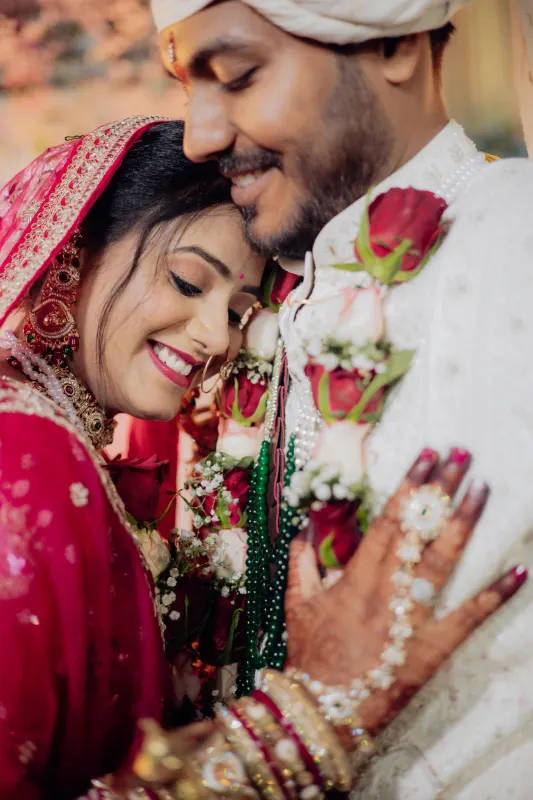Smiling Indian bride in red dupatta rests her head against the groom's rose garland